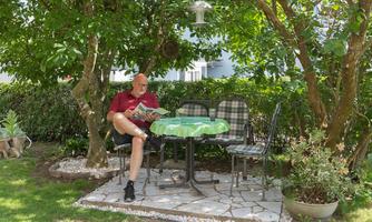 A man is sitting at a table in the garden reading a newspaper. Trees are growing around him and there is a relaxed, green atmosphere.