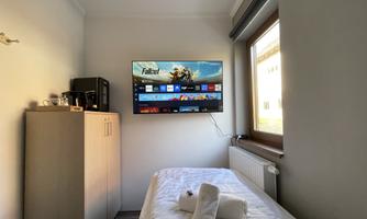 A simple room with a single bed and a television on the wall. Next to the bed is a small wardrobe and a window lets in natural light.