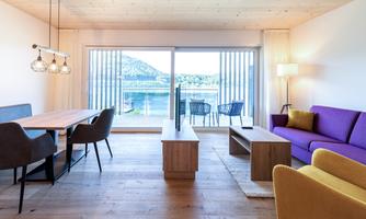 A modern living room with a purple sofa and a dining area. Large windows offer a view of the water and the surrounding landscape.