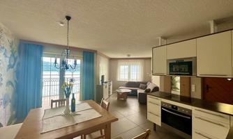 A modern living kitchen with a dining table and comfortable seating. Large windows provide plenty of light and a view outside.