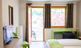 A modern room with a large window and access to a balcony. It is brightly lit and features simple furniture.