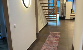 A modern hallway with a wooden staircase and bright light. The floor is covered with a colorful rug, and the walls are painted in neutral colors.