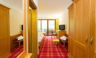 A bright hotel room with wooden furniture and a red carpet. There is a television, a table, and a balcony with a view outside.
