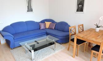 A modern living room with a blue couch and a glass table. Next to it is a wooden table with chairs and a plant.