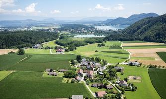 A picturesque landscape with green fields and a small village. In the background, mountains and a lake can be seen.