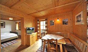 A cozy room with wooden interior and a dining table. In the background, there is a television and large windows for plenty of daylight.
