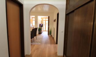 A bright hallway with wooden flooring and an open access to a living area. Doors can be seen on the left side and a dining area in the background.