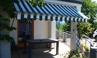 A daylight area with a striped sunshade and a table. The surroundings are green and inviting.