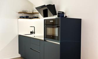 A modern kitchen with an elegant dark kitchen unit and a built-in oven. The room has bright walls and a warm wooden floor.