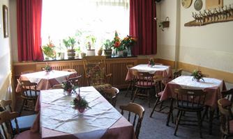 A cozy restaurant with tables and chairs. The windows are bright and surrounded by plants.
