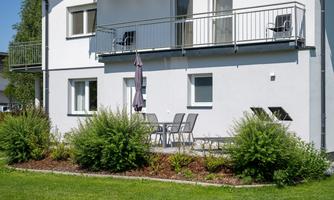 A modern house with a balcony and a well-maintained garden. There are garden furniture and a parasol on the terrace.