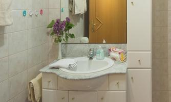 A bright bathroom with a sink and a large mirror. Next to the sink are a flower vase and various personal care products.