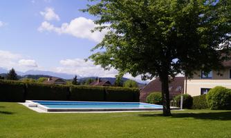 A peaceful garden with a swimming pool and a large green lawn. In the background, there are trees and a bright blue sky.