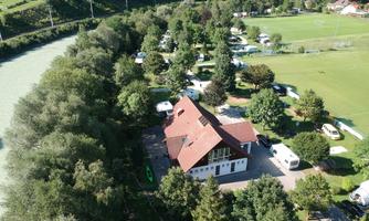 A spacious campground with a large house and many trees. In the background, there are motorhomes and a sports field.