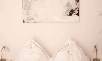 A cozy bedroom with a large white bed and two pillows arranged in a heart shape. An abstract painting in neutral colors hangs on the wall.