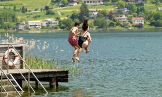 Two people jump together from a pier into a clear lake. In the background, green hills and houses can be seen.