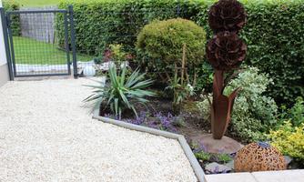 A well-maintained garden with a gravel path, various plants, and a modern sculpture. In the background, a garden fence is visible, surrounded by green hedges.