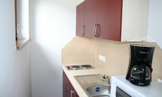 A small kitchenette with red cabinets and a bright countertop. There is a kettle, a microwave, and cooktops.