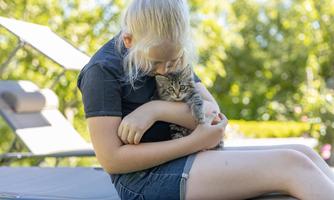 A girl is sitting on a lounger, holding a small kitten in her arms. It is a sunny day with a lot of green in the background.