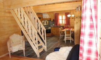 A cozy wooden house interior with a staircase, a dining area, and a modern kitchen. The windows are decorated with red curtains, and there is soft seating.