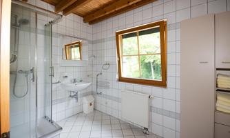 A bright bathroom with white tiles and a window that lets in plenty of light. There is a shower, a sink, and a cabinet with towels.