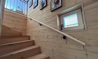 A wooden staircase with a railing leads up. Pictures hang on the wall, and a small window lets light in.