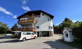 A multi-story building with balconies and a white delivery van in front of it. The sky is blue with some clouds and there is plenty of greenery in the surroundings.