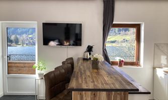 A modern living space with a wooden bar, comfortable chairs, and a television on the wall. Through the window, there is a beautiful view of nature.