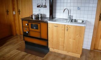 A small, rustic kitchen with an old stove and a stainless steel sink. The walls are made of wood and the floor is covered with laminate.