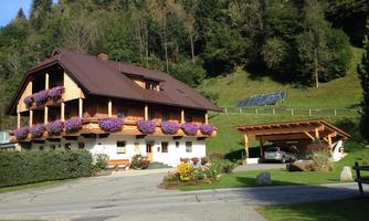 A picturesque house with blooming flower boxes and a carport. In the background, gentle hills and trees can be seen.
