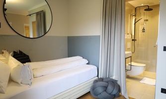 A modern bedroom with a cozy bed and a large mirror. In the background is a bright bathroom with a shower.