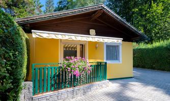A cozy holiday home in lively yellow with a small balcony. Flowers in a pot beautify the entrance area and are surrounded by green shrubs.