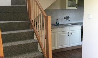 A bright interior with a staircase and a small kitchen. The floor is made of wood, and the walls are kept neutral.