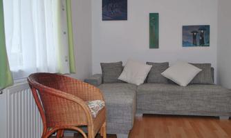 A cozy living space with a gray sofa and two cushions. A wicker chair and some pictures on the wall complete the scene.