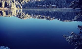 A tranquil lake with reflective water and snow-covered trees. In the background, the snow-capped mountains and a lodge can be seen.
