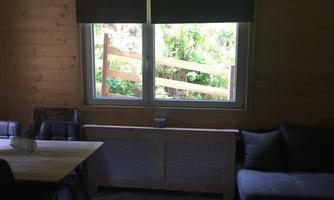 A cozy room with wooden paneling, a table, and a couch. Through the window, a green landscape with a wooden fence can be seen.