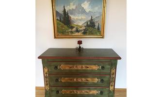 A decorative chest of drawers with golden embellishments and a green hue is in the foreground. Above it hangs a landscape painting with mountains and trees.