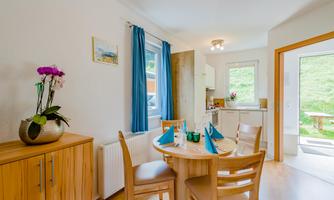 A cozy dining area with a wooden table and chairs. The room is bright and friendly, with an open kitchen and access to the terrace.