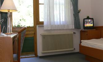 A simple room with a bed, a desk, and a television. Lots of light streams through the windows with curtains.