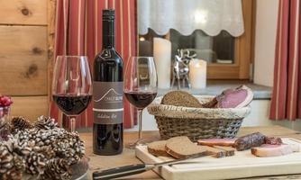 A bottle of red wine with two glasses is on a table. Next to it lies a basket of bread and a selection of sausage and cheese.