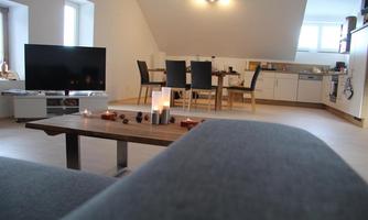 A modern living room with a gray sofa and a television. In the background, there is a dining table and an open kitchen.