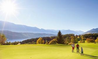 A sunny golf course landscape with a view of mountains and a lake. Three people are standing on the green and enjoying the view.