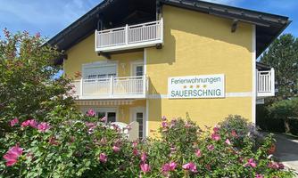 A sun-yellow painted building with two balconies and a sign that says "Ferienwohnungen Sauerschnig." Colorful plants are blooming in front of the house.