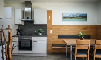 A modern kitchen with light furniture and wooden paneling. A dining table with chairs and a flower on the table is also visible.