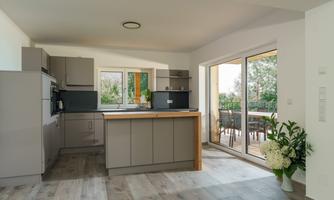 A modern kitchen with gray cabinets and a central cooking block. Large windows offer a view of the terrace and the garden.
