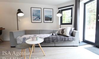 A modern living room with a gray sofa and a small table. The large windows let in plenty of natural light and there are artistic paintings on the wall.