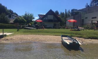 A quiet lake with a boat in the foreground and a meadow in the background. In the background, a house and sunshades are visible.