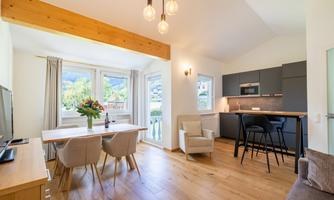 A modern living room with wooden flooring, table, and chairs. The large window front ensures plenty of natural light and a view of the greenery.