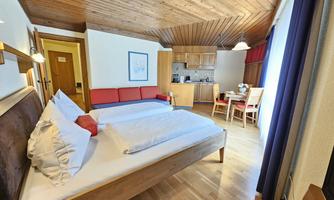 A cozy room with a double bed and an additional single bed. The furnishings are made of wood, featuring a small kitchen and a dining table.