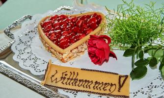 A heart-shaped cake with red fruits and a beautiful decoration of roses. Next to it lies a board with the inscription "Welcome!".